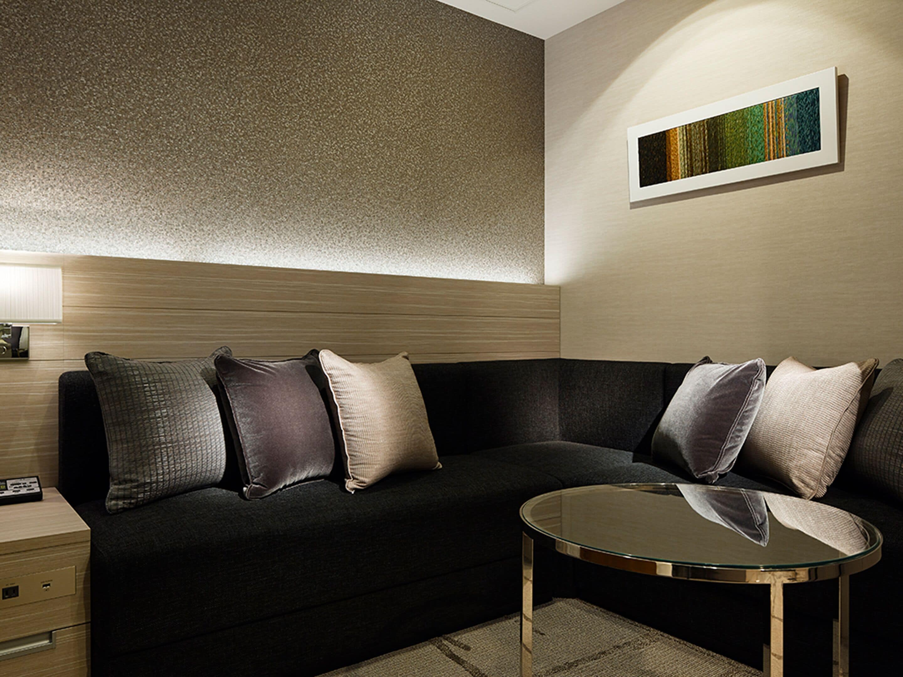 A modern hotel room with a dark-colored sofa, a glass coffee table, a lamp, and a painting on the wall.