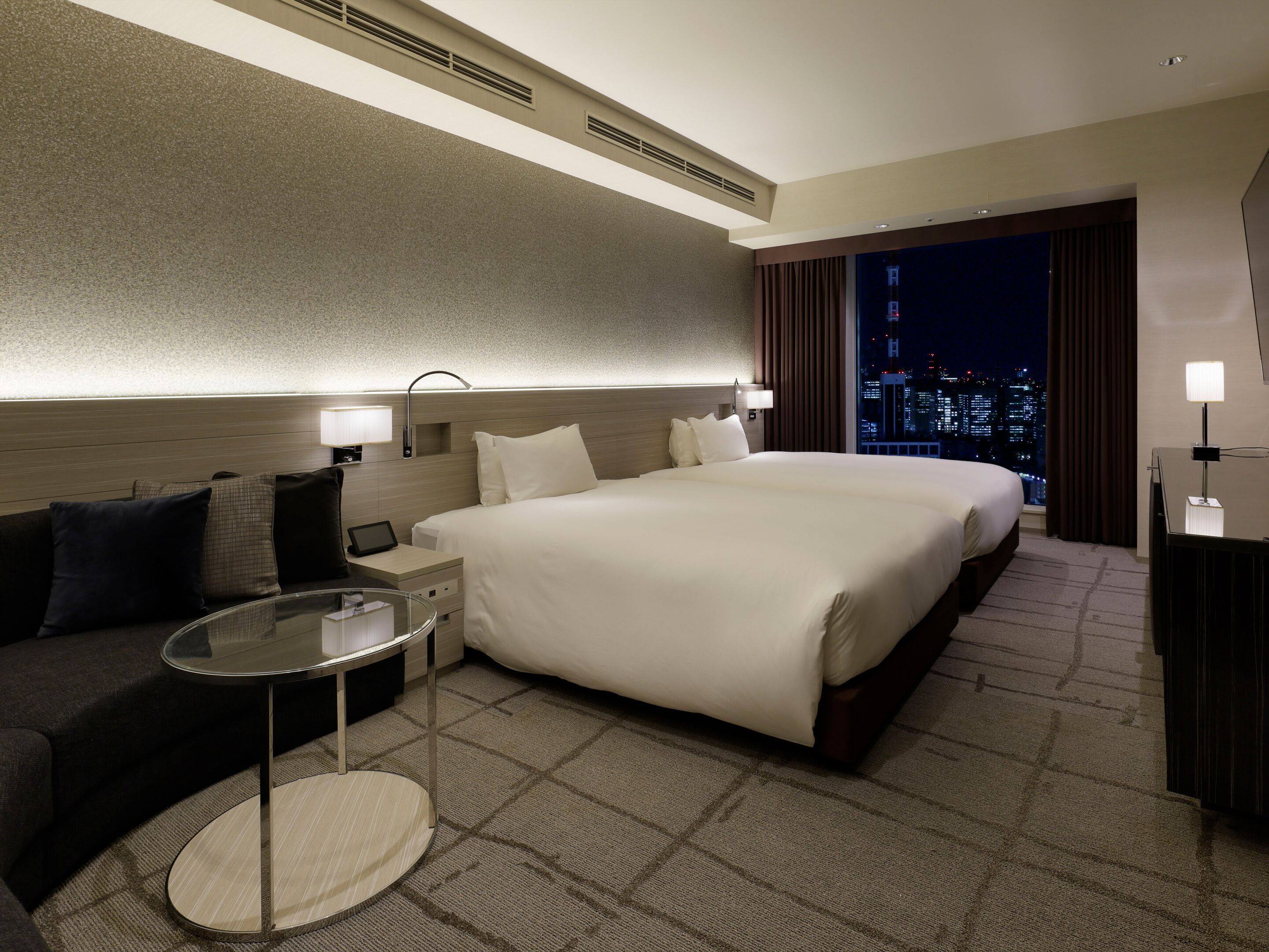 Habitación de hotel con una cama grande, un sofá, una mesita y vista a la ciudad de noche.
