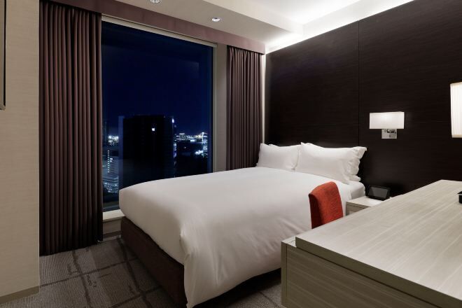 A hotel room with a large bed, a nightstand, a desk, and a view of the city at night.