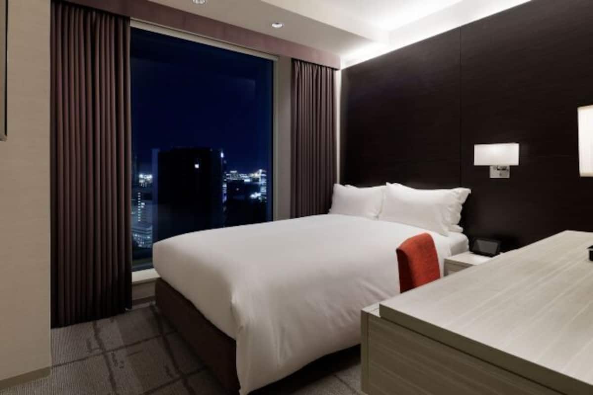 A hotel room with a large bed, a nightstand, a desk, and a view of the city at night.