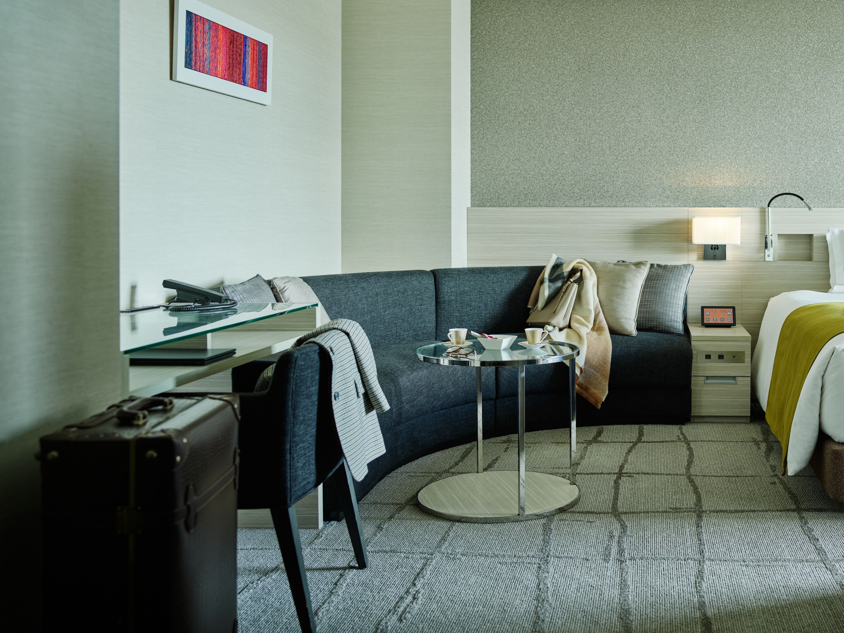 A modern hotel room with a curved sofa, a small round table, a glass desk, a suitcase, and a bed with a yellow blanket.