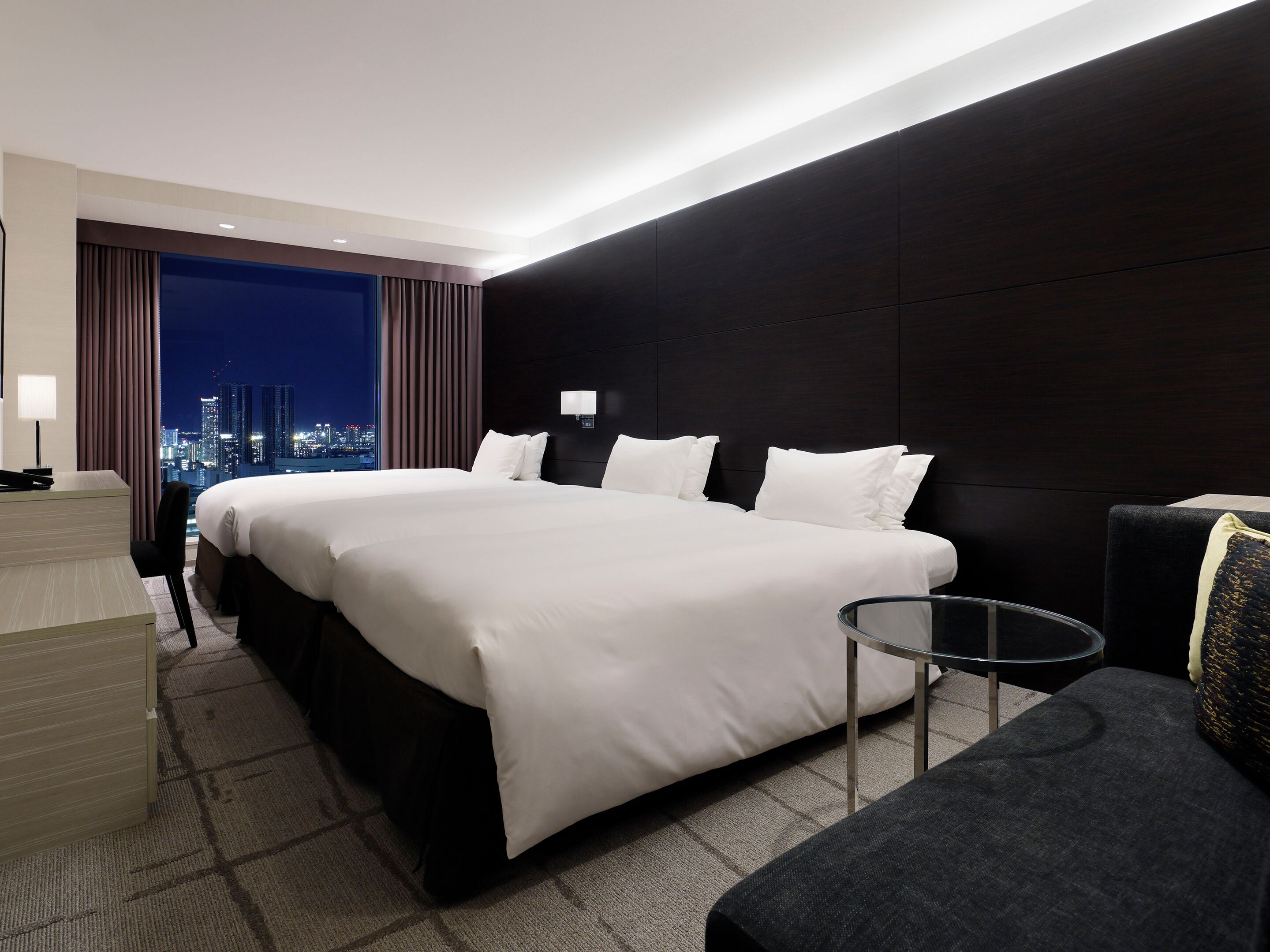 A modern hotel room with a large bed, a desk, a chair, and a view of the cityscape at night.
