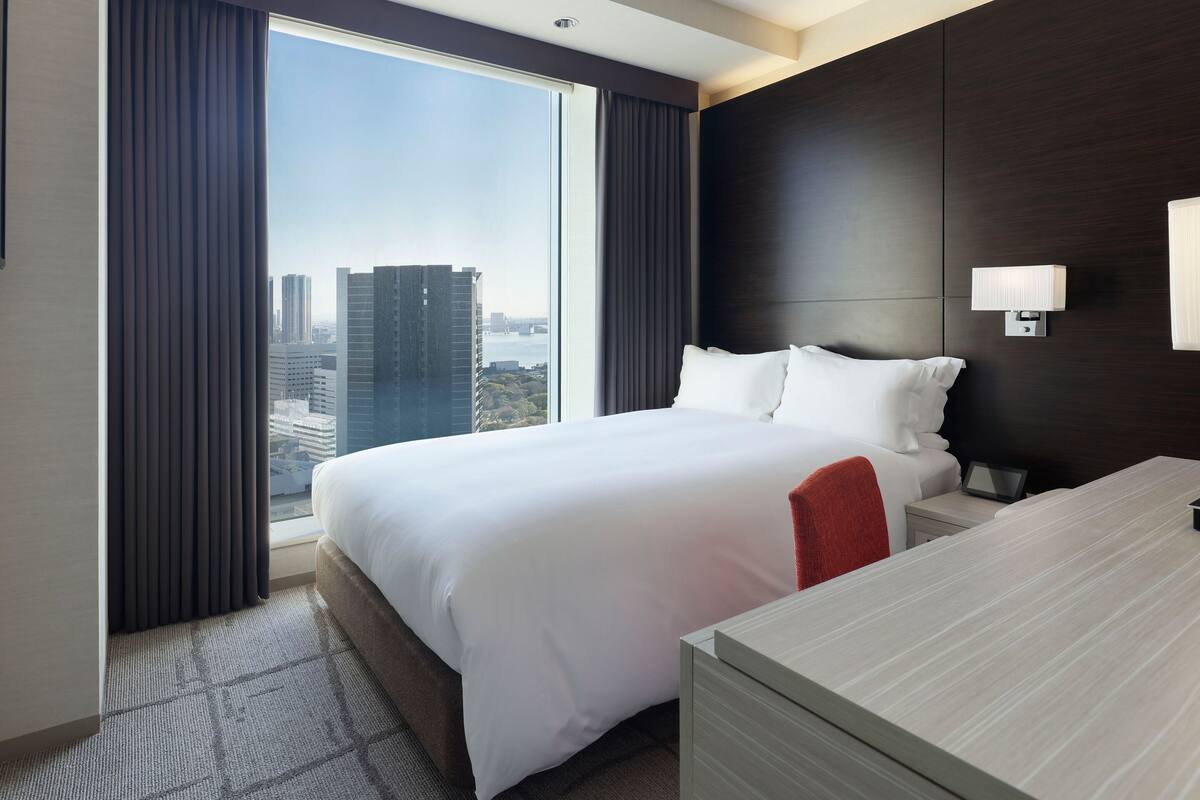 Une chambre d’hôtel avec un grand lit, un bureau, une table de chevet, une lampe et une vue sur le paysage urbain par la fenêtre.