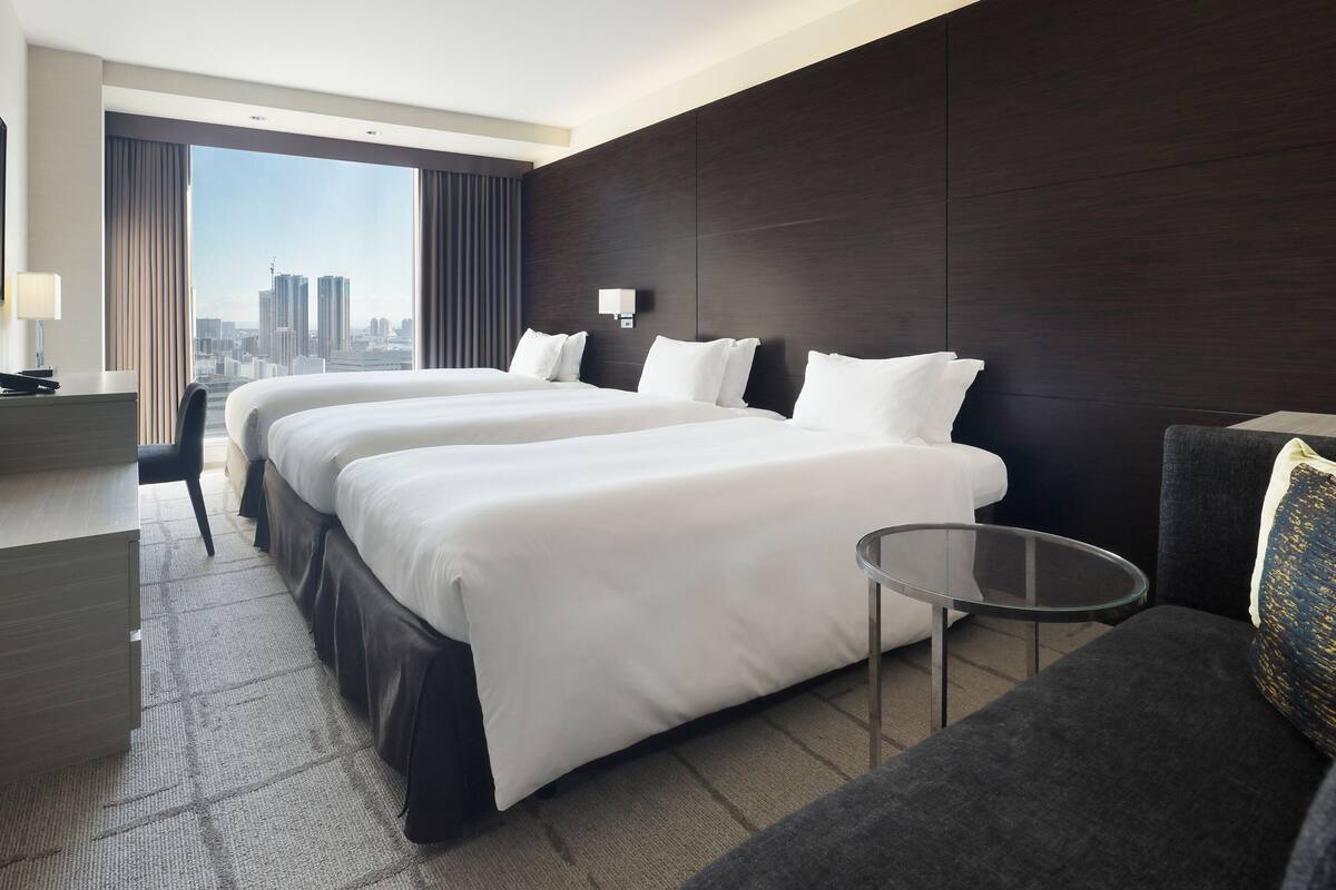 Une chambre d’hôtel avec deux lits, un bureau, une chaise, une petite table et une grande fenêtre donnant sur la ville.