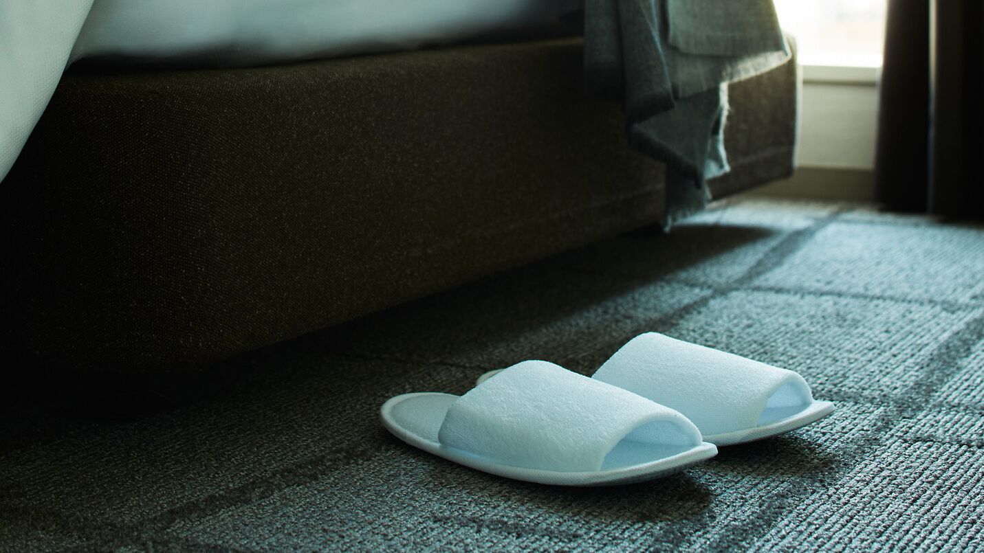 A pair of white slippers on a textured carpet near a dark-colored sofa.