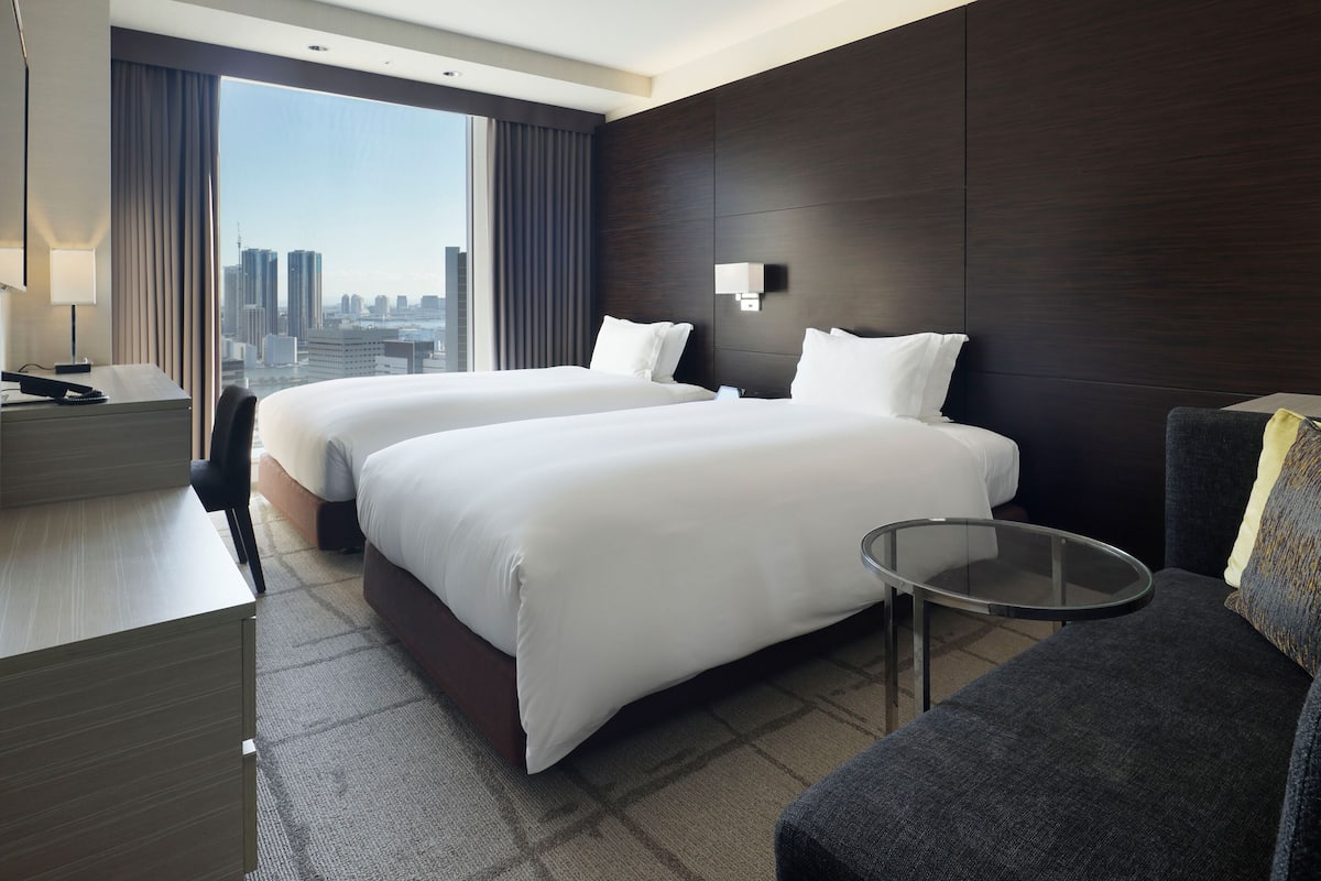 Une chambre d’hôtel avec deux lits, un bureau, une chaise, une petite table et une vue sur le paysage urbain.