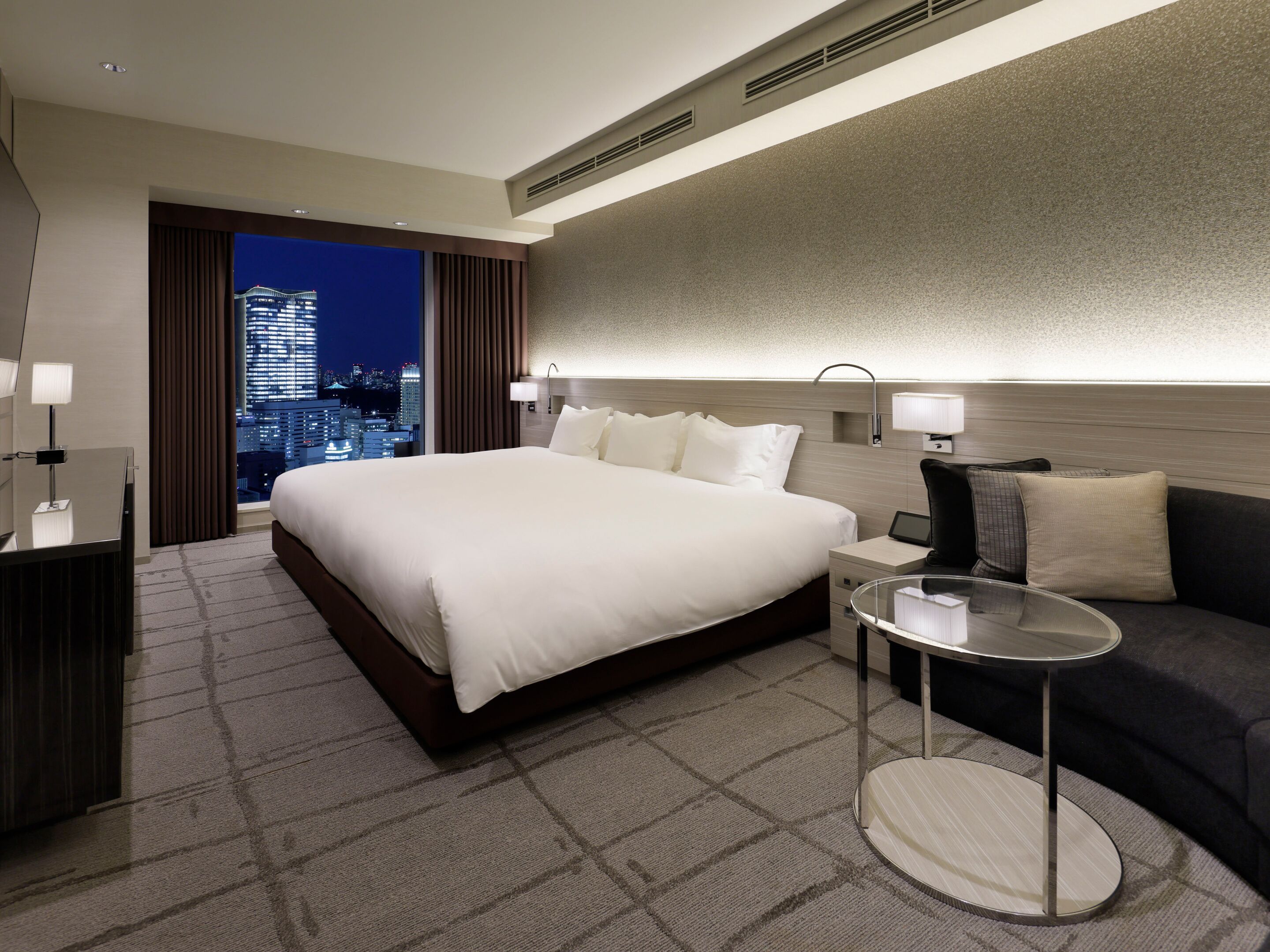 Habitación de hotel con una cama grande, una mesita de noche, una mesa redonda de vidrio, un sofá y vistas a la ciudad.