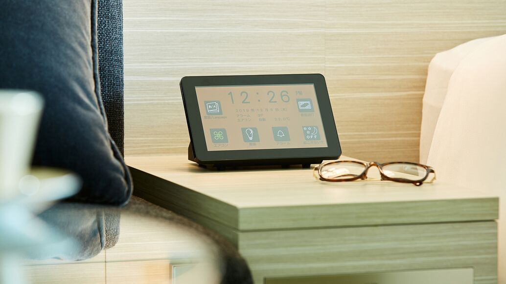 A bedside table with a digital clock displaying 12:26, a pair of glasses, and a pillow.