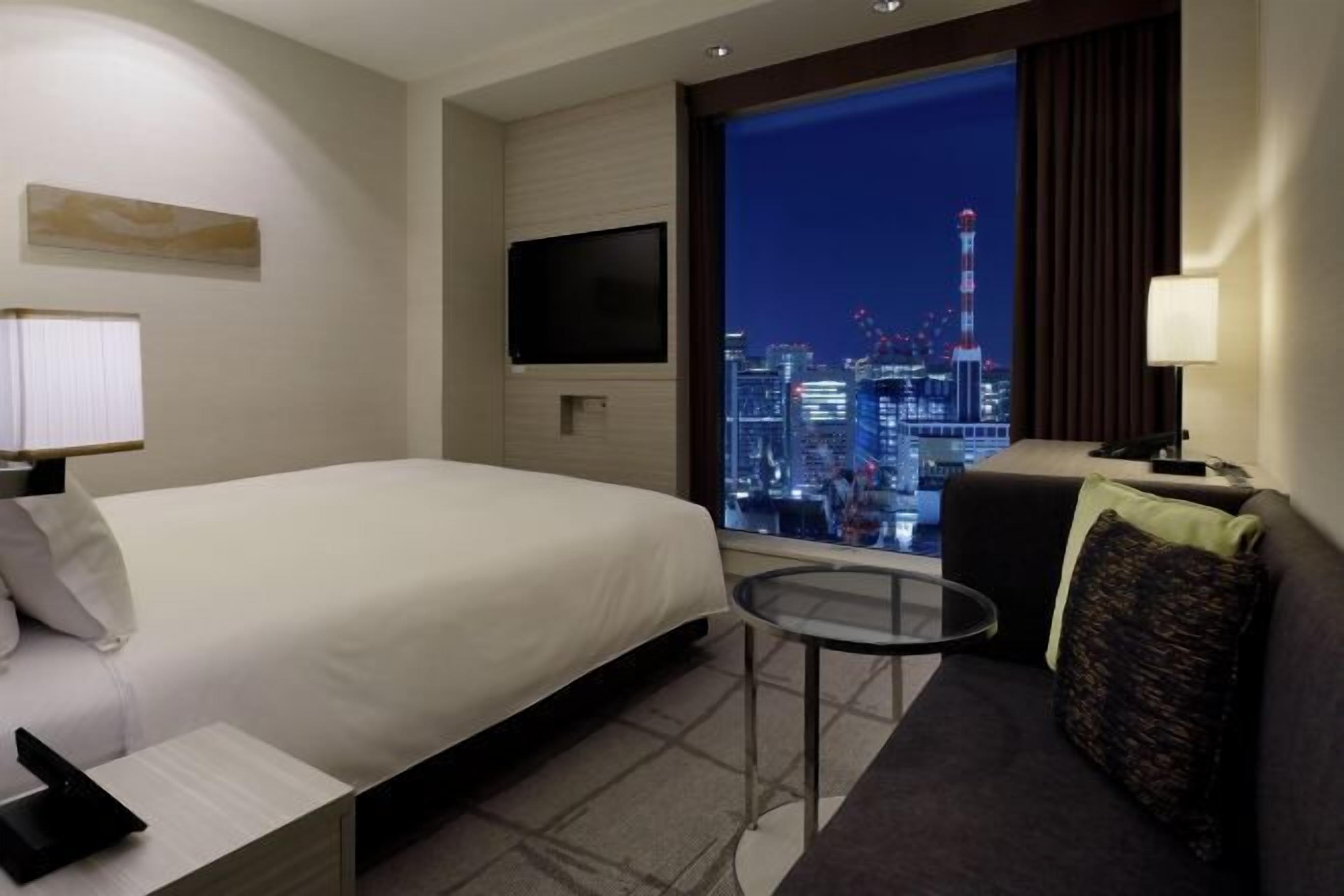 A hotel room with a bed, a small table, a sofa, a TV, and a view of a cityscape at night.