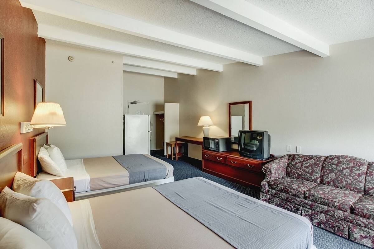 Quarto de hotel com duas camas, uma escrivaninha com computador e luminária, televisão e sofá floral.