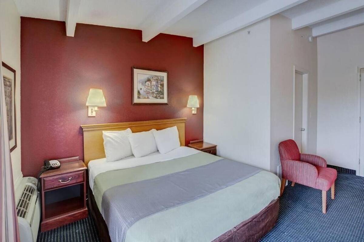 Quarto de hotel com uma cama, duas luminárias de cabeceira, uma cadeira vermelha e um quadro pendurado na parede.