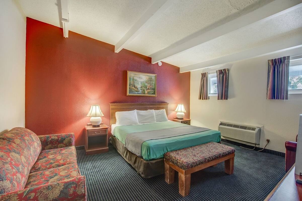 Quarto de hotel com cama, duas mesas de cabeceira com luminárias, um banco, radiador e janela com cortinas.