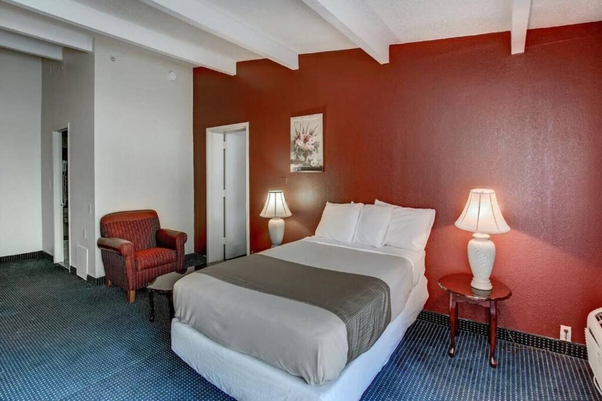 Quarto de hotel com cama, duas luminárias de cabeceira, uma cadeira e uma porta.