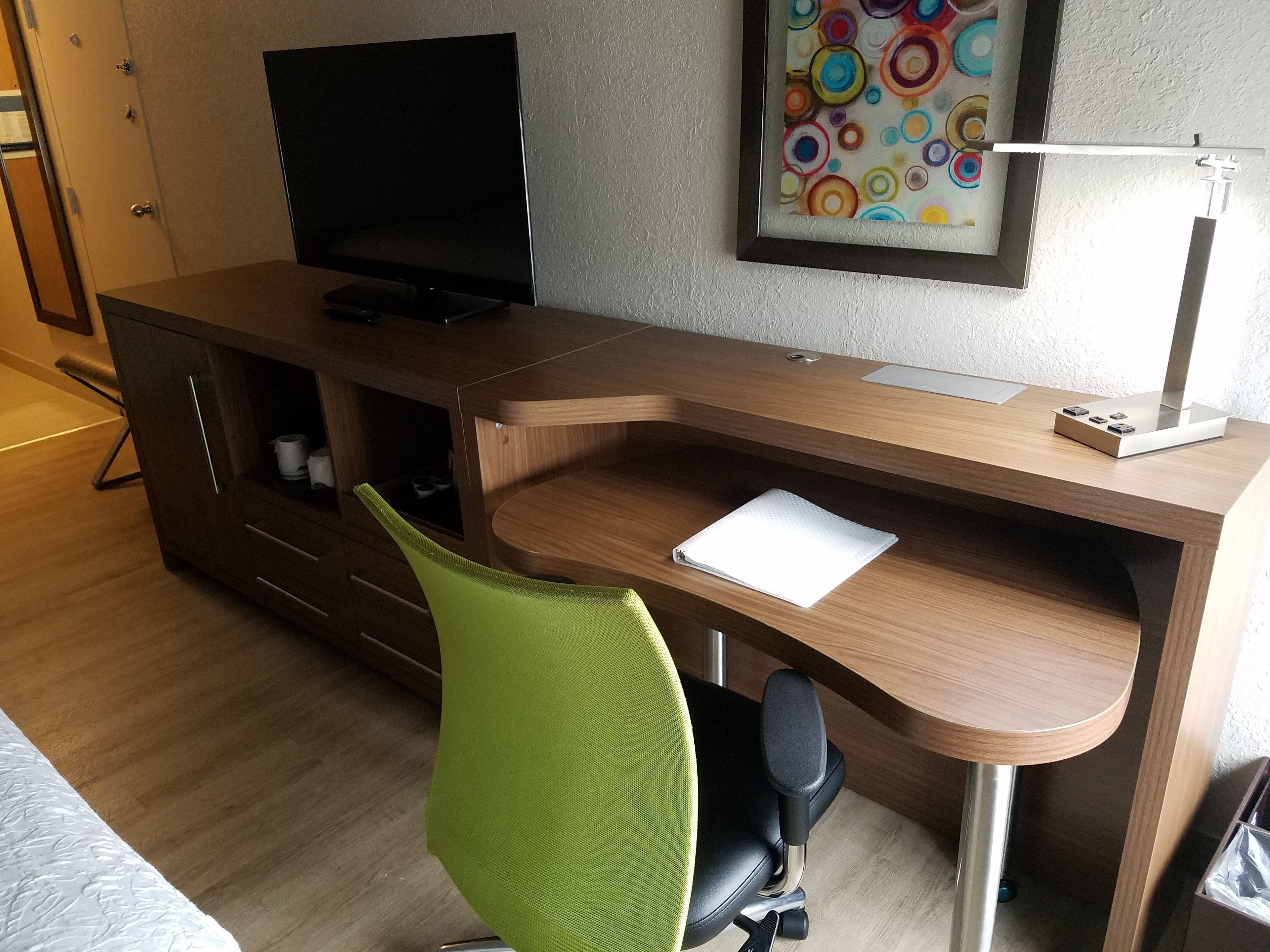 A modern office space with a wooden desk, a green office chair, a television on a cabinet, and a colorful abstract painting on the wall.