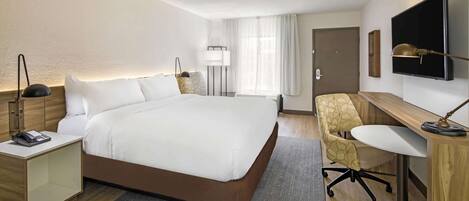 Habitación de hotel con una cama grande, mesita de noche con un teléfono, escritorio con una silla, televisor montado en la pared y una puerta.