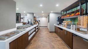 Un comptoir-buffet moderne, doté de plusieurs distributeurs de nourriture, d’une machine à café et d’un présentoir réfrigéré contenant des fruits et des céréales.