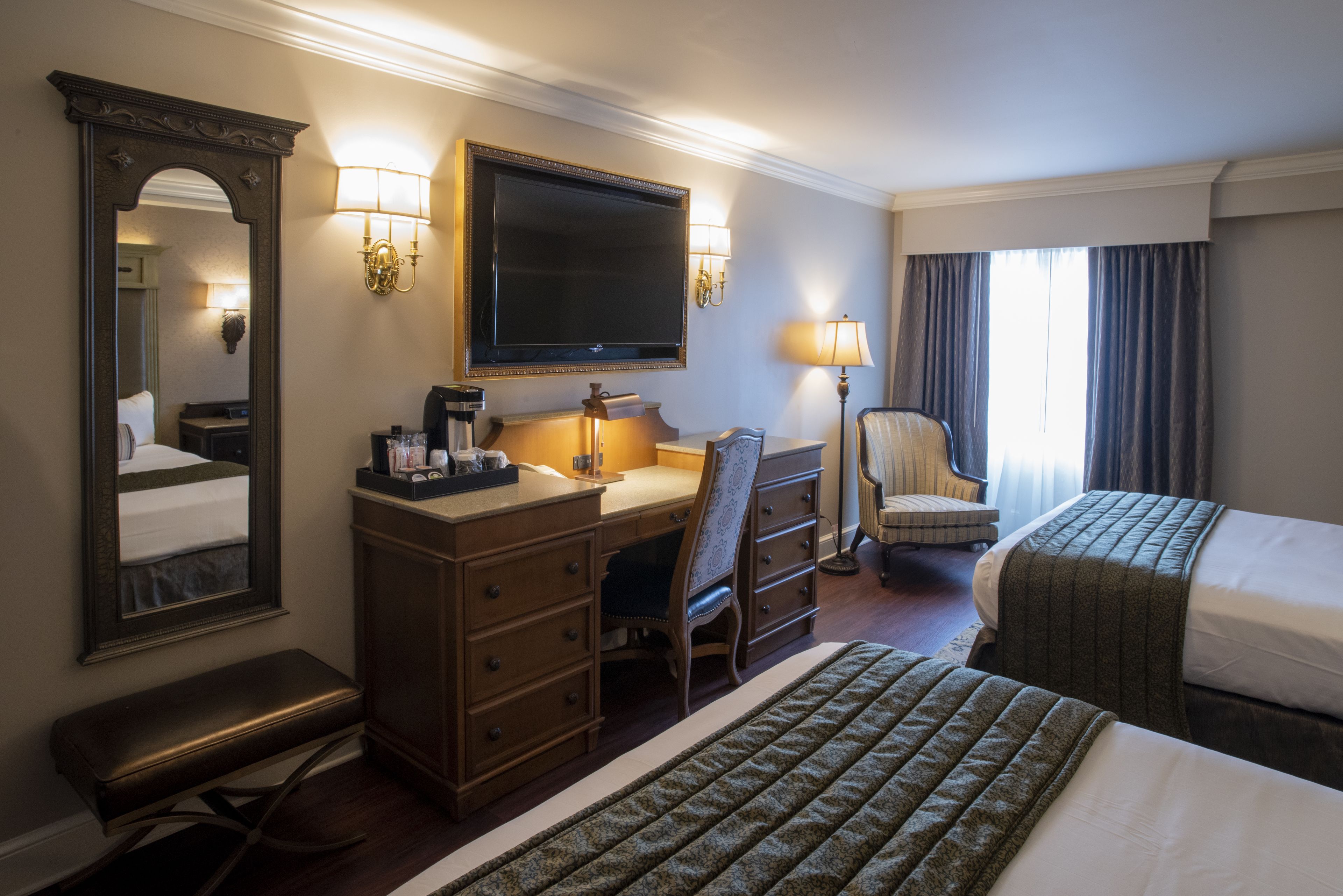 A hotel room with a large bed, a smaller bed, a desk with a chair, a mirror, a flat-screen TV, and two wall-mounted lamps.