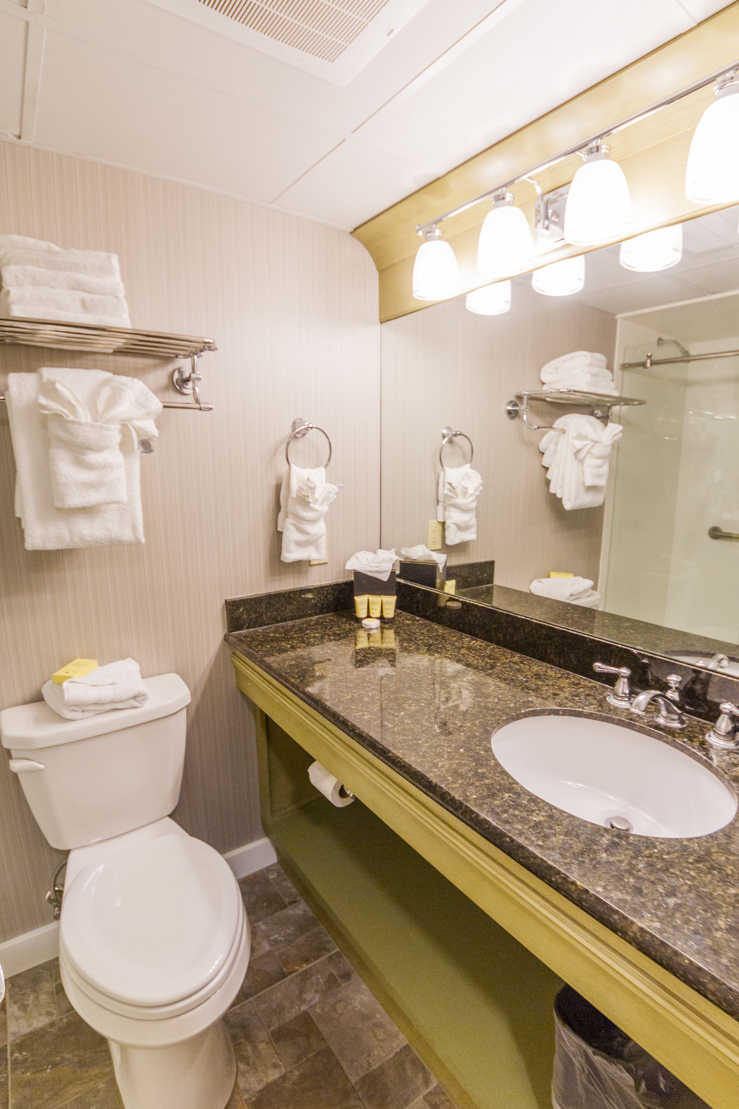 A bathroom with a toilet, sink, and mirror. There are towels hanging on the wall and a shelf with towels.