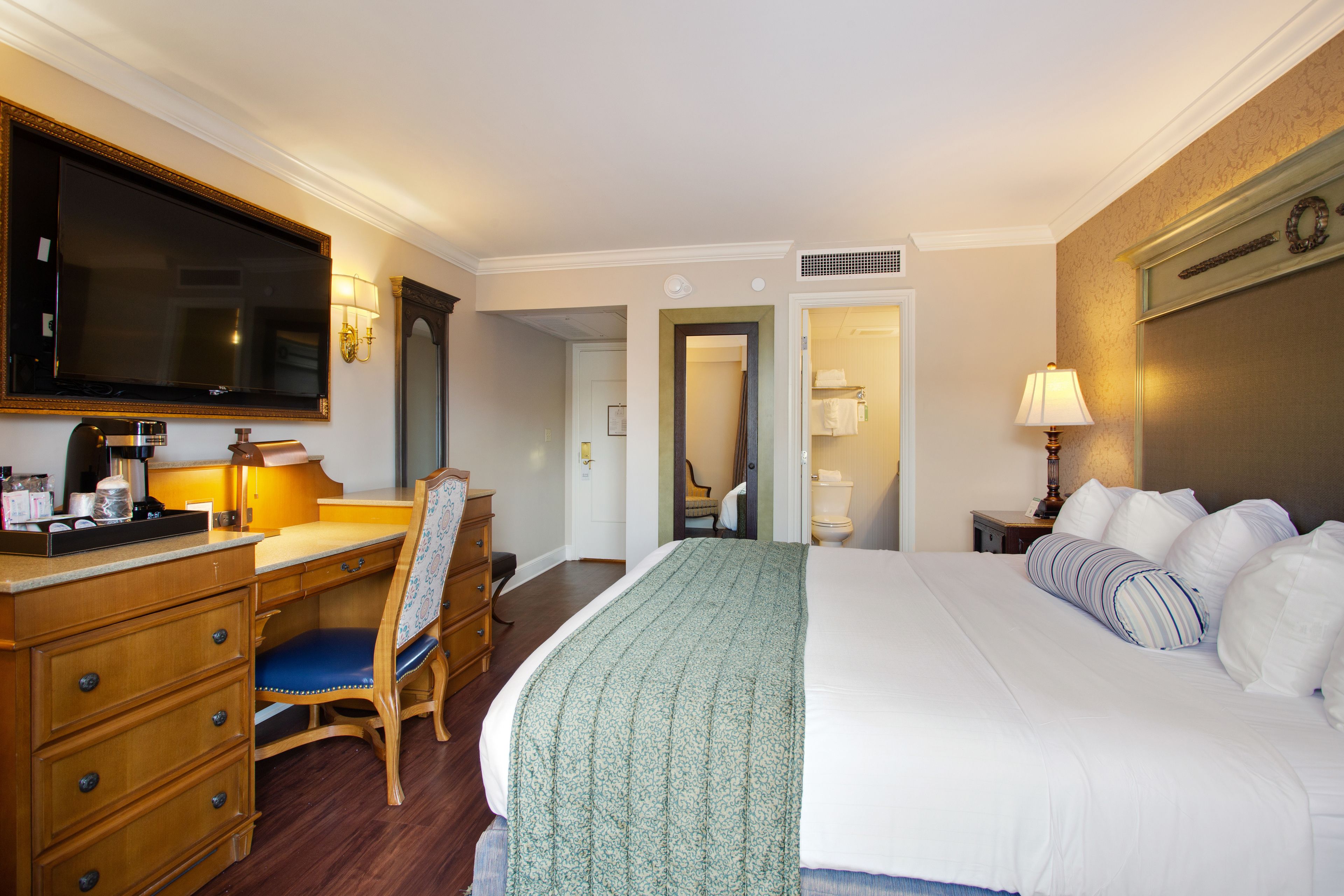 A hotel room with a large bed, a desk with a chair, a television, and a bathroom visible through an open door.