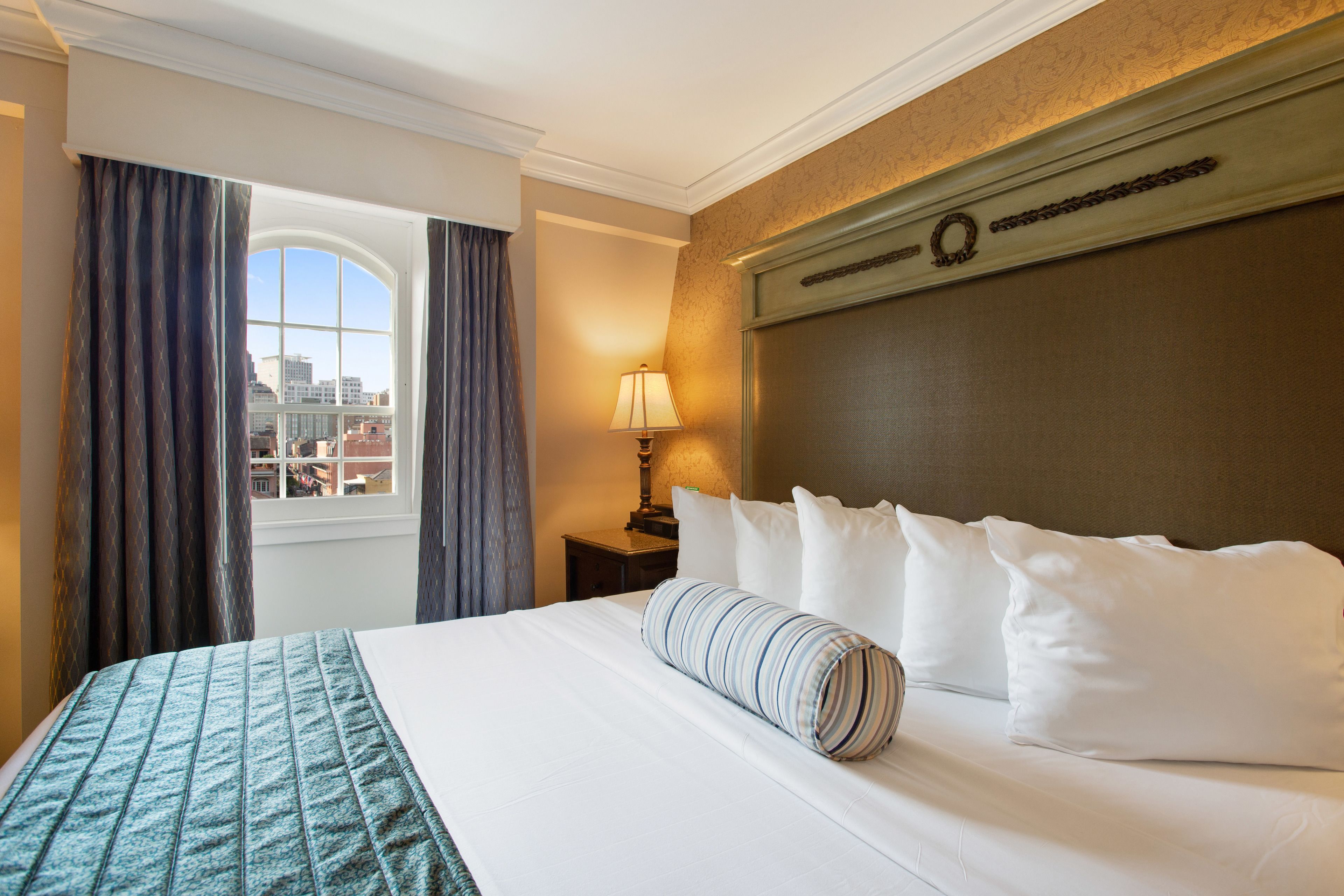 Quarto de hotel com uma cama grande, janela com cortinas, mesa de cabeceira com abajur e vista da cidade.