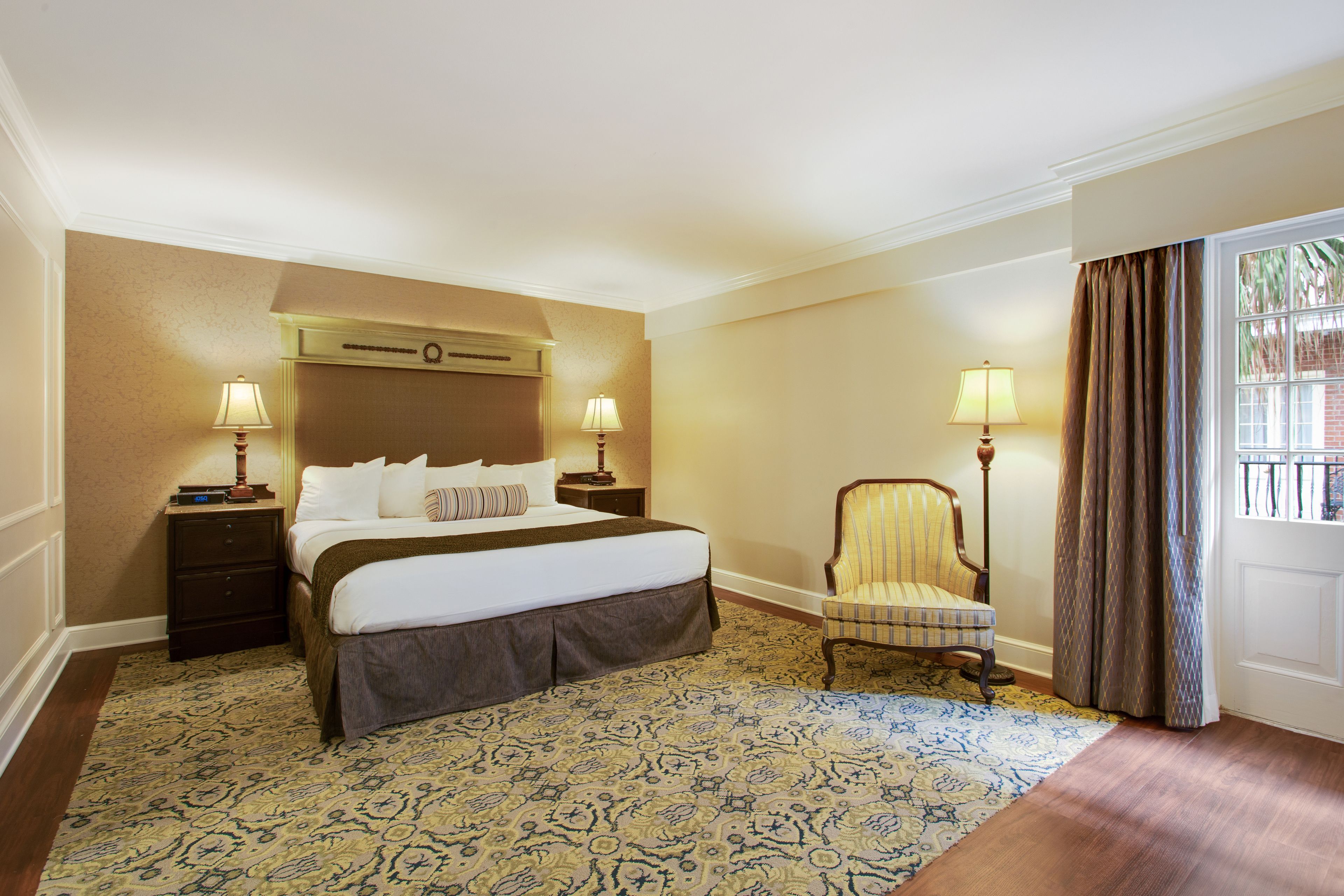 Habitación de hotel con una cama grande, dos mesitas de noche con lámparas, una silla, alfombra estampada y una ventana con cortinas.