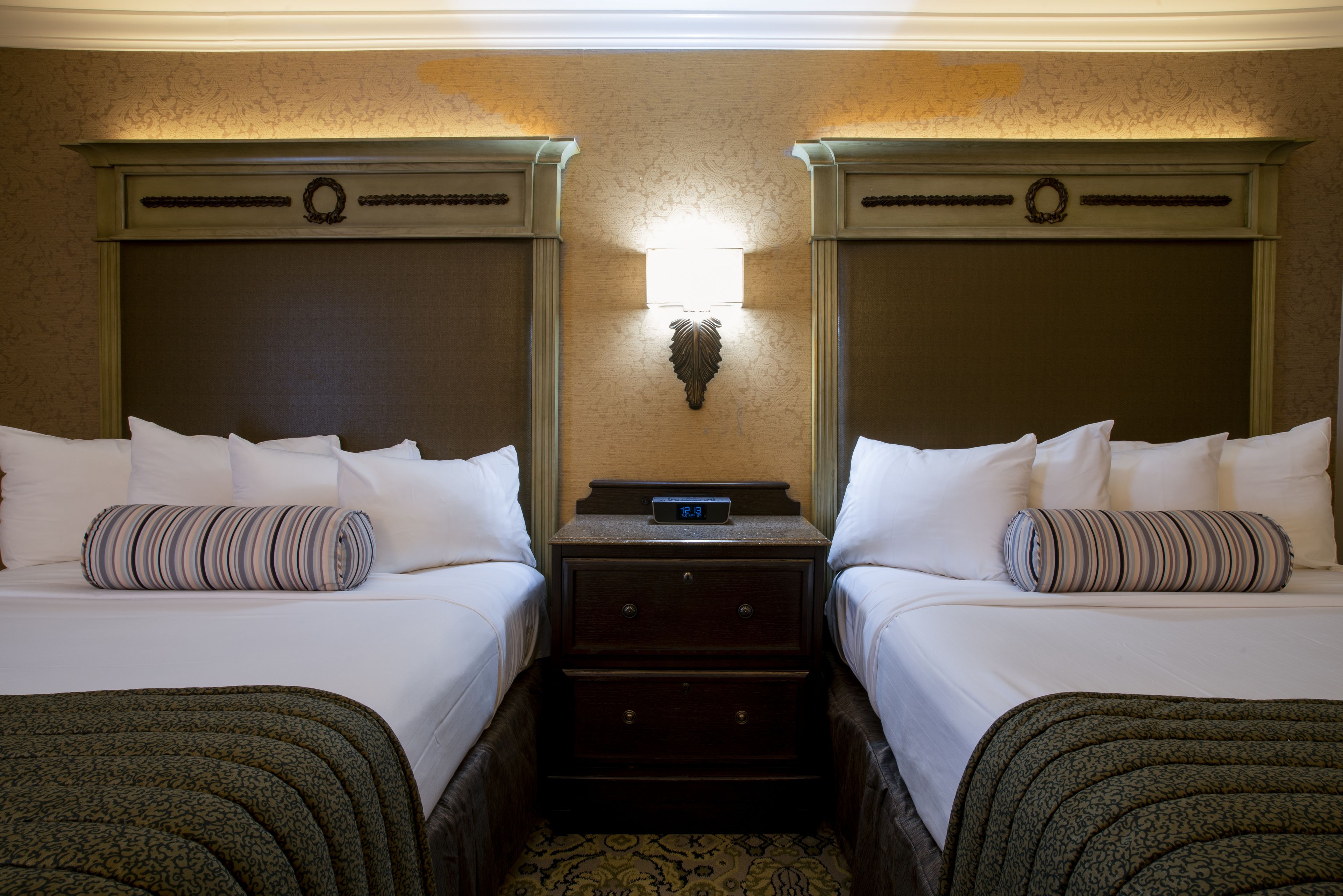 A hotel room with two beds, a nightstand with a clock, and a wall-mounted light fixture.