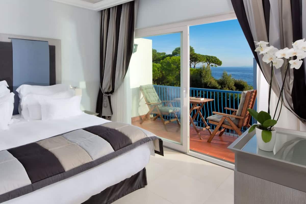 Une chambre d’hôtel avec un lit, un balcon donnant sur la mer et une petite table avec des chaises.