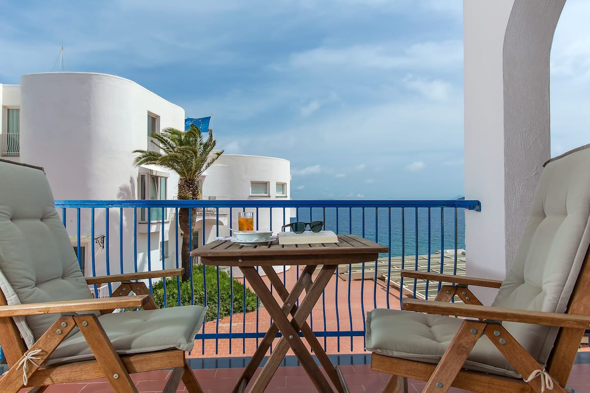 Un balcon avec deux chaises en bois, une petite table sur laquelle se trouvent un livre, des lunettes de soleil et une boisson. La vue comprend un bâtiment blanc et la mer.