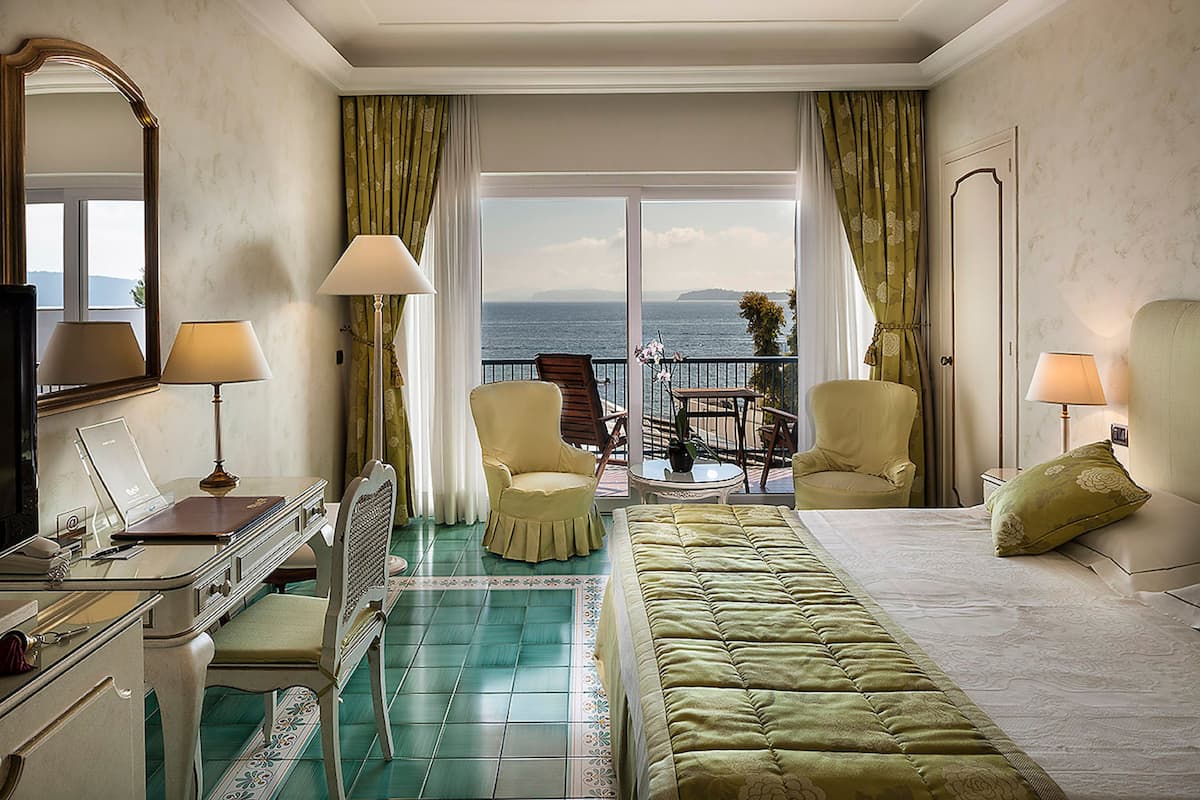 Une chambre d’hôtel avec un lit, deux chaises, un bureau et un ordinateur portable. Elle dispose d’un balcon avec vue sur la mer.