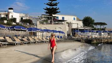 Una playa con tumbonas y sombrillas, una mujer en traje de baño caminando por la orilla, y edificios al fondo.