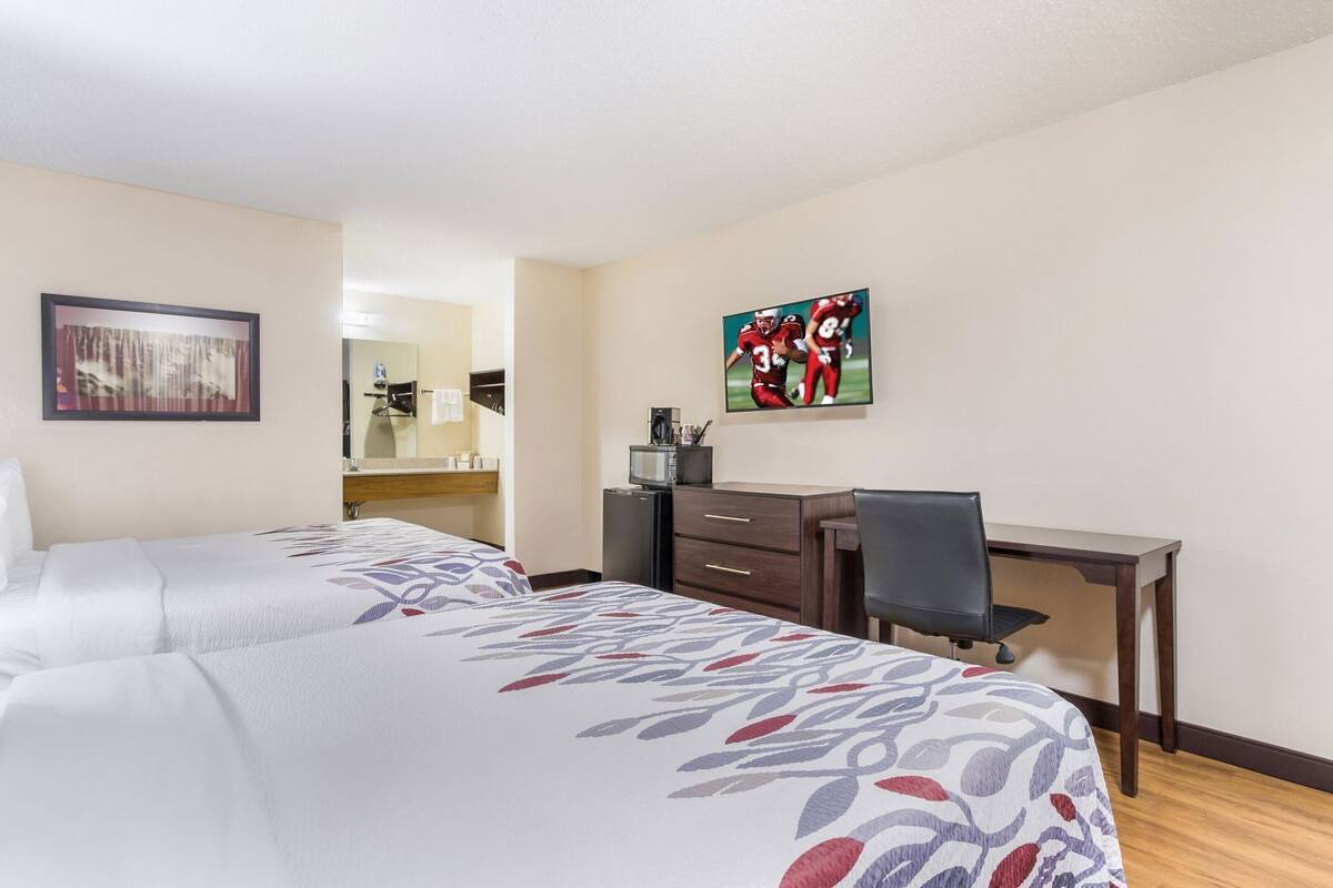 A hotel room with two beds, a desk, a chair, a TV, and a painting on the wall.