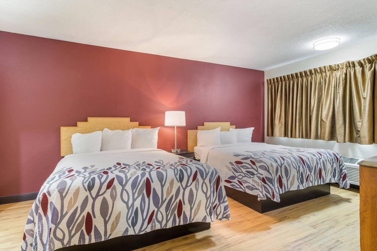 A hotel room with two beds, a nightstand, a lamp, and a window with curtains.