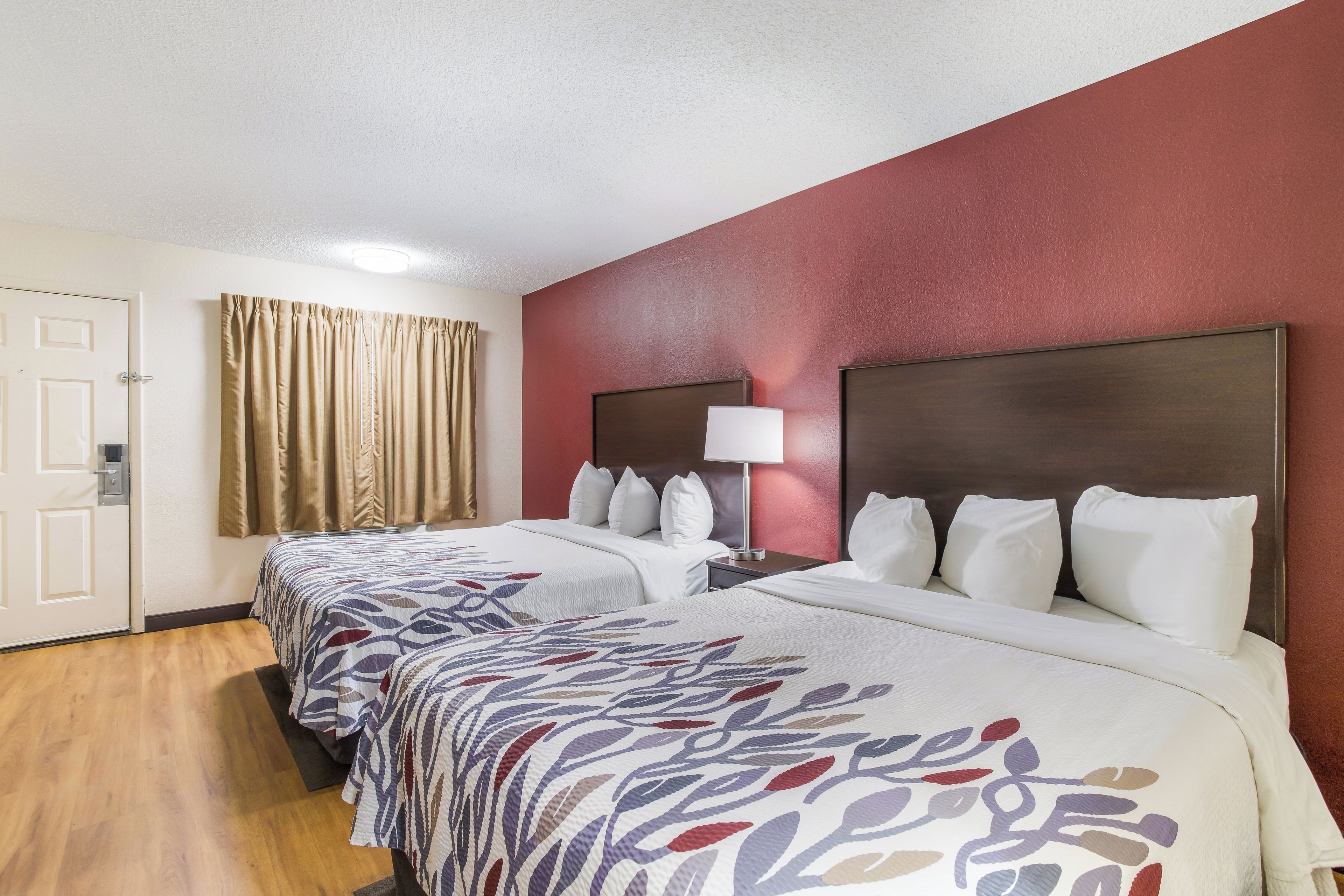 A hotel room with two beds, a nightstand with a lamp, a door, and a window with curtains.