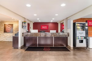 Zona de recepción de hotel con mostrador de mármol, estación de registro digital, pared roja con señal de Wi-Fi y una máquina expendedora.