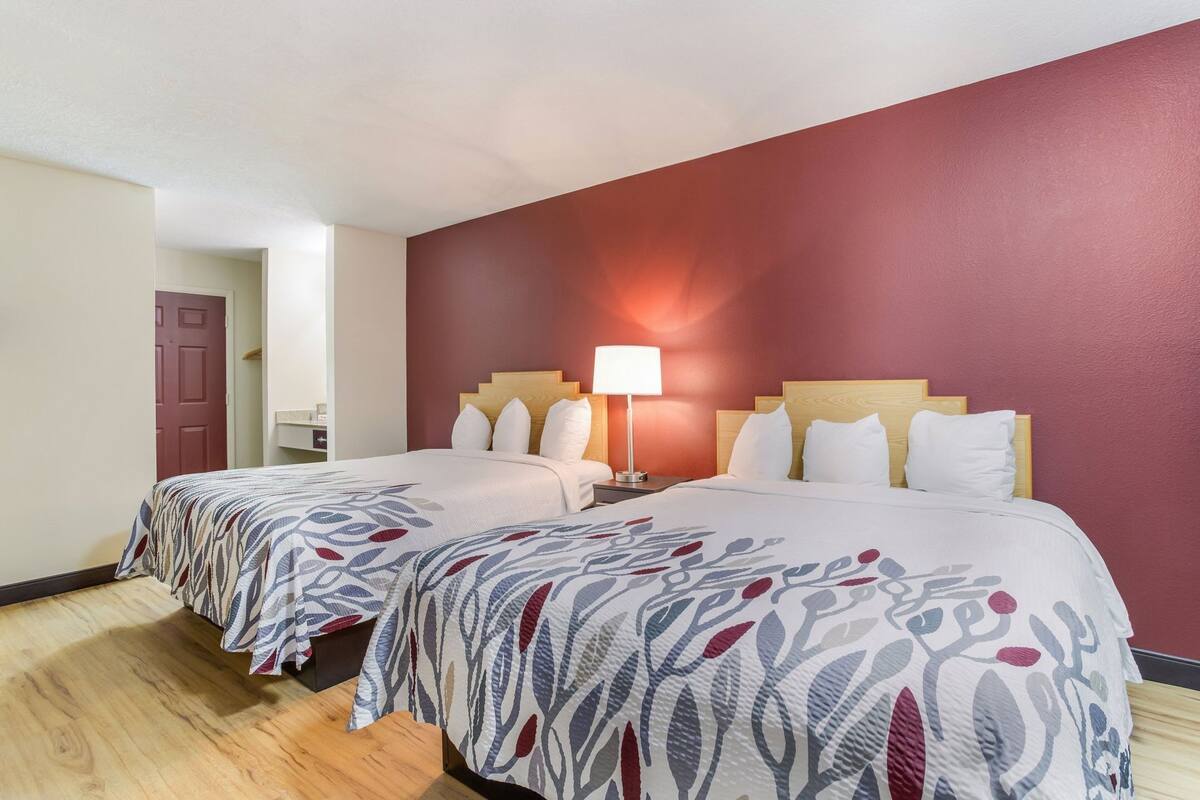 A hotel room with two beds, a nightstand, a lamp, and a red accent wall.
