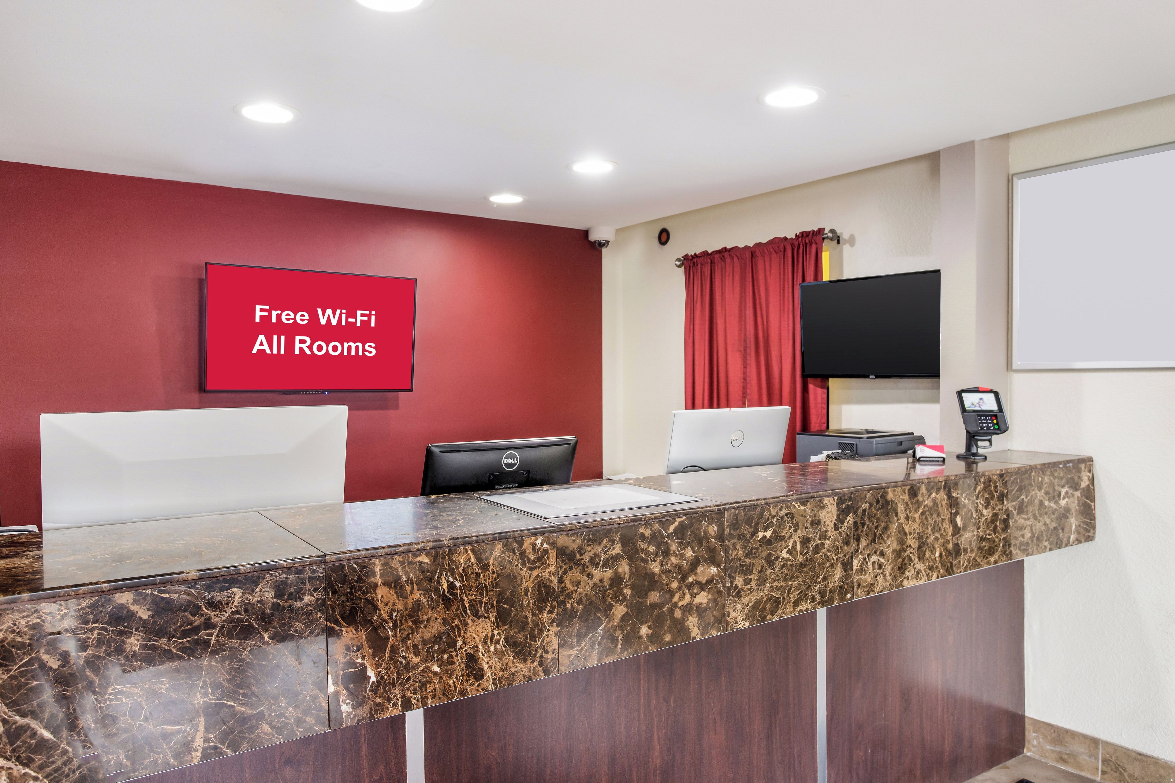 Un comptoir d’accueil avec un plateau en marbre, un mur rouge orné d’un panneau indiquant « Wi-Fi gratuit dans toutes les chambres », un téléviseur à écran plat et un ordinateur Dell.