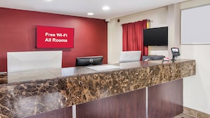 A reception desk with a marble top, a red wall with a 'Free Wi-Fi All Rooms' sign, a flat-screen TV, and a Dell computer.