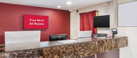 Una recepción con encimera de mármol, una pared roja con un cartel que dice 'Wi-Fi Gratis en Todas las Habitaciones', un televisor de pantalla plana y un ordenador Dell.