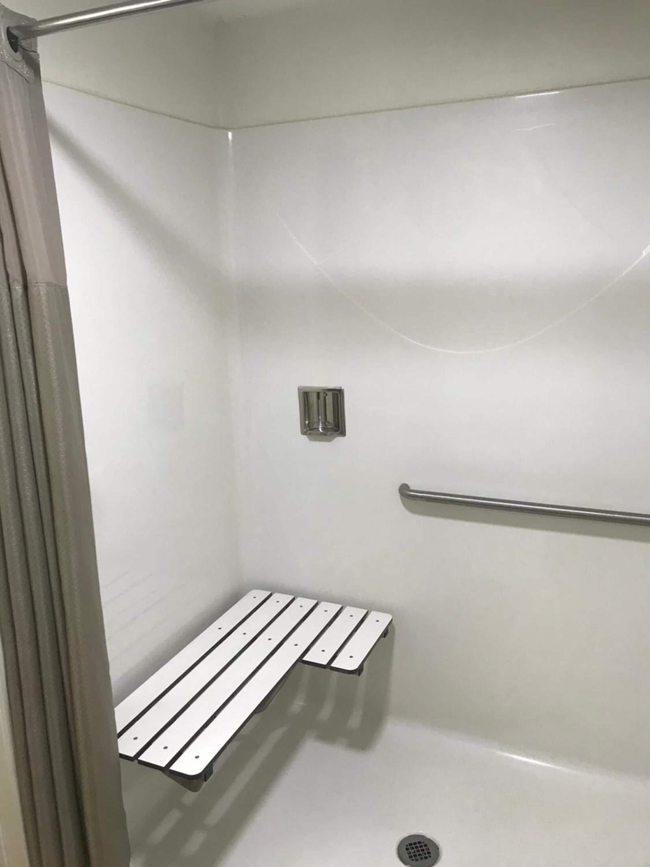 Ducha con pared de azulejos blancos, barra de metal de apoyo, asiento en la ducha y desagüe.