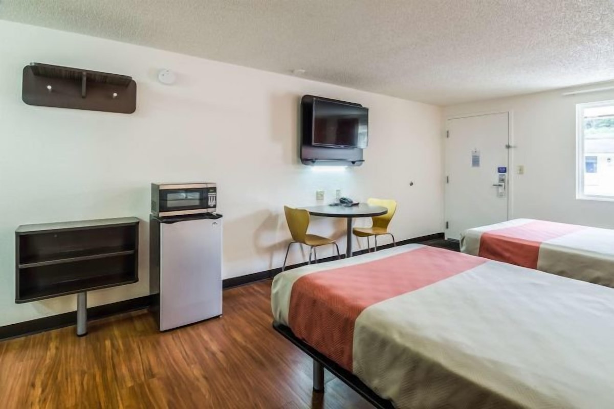 A hotel room with two beds, a small desk with a computer, a TV mounted on the wall, and a window providing natural light.