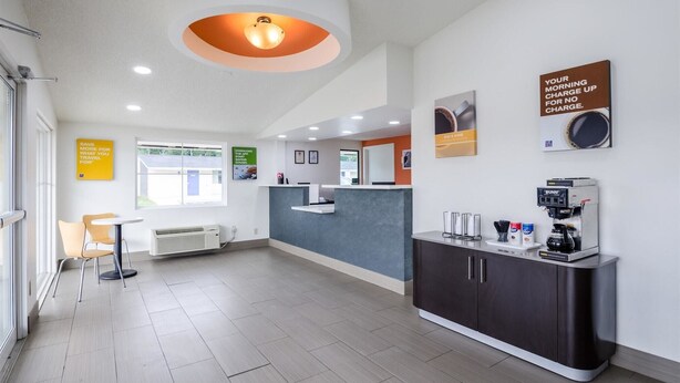 A modern hotel lobby with a reception desk, a coffee machine, and a seating area with chairs.