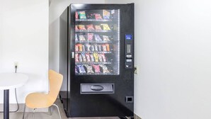 A vending machine with various snacks and drinks, located in a room with a white table and a yellow chair.