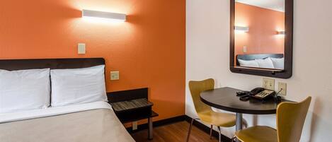 Habitación de hotel con cama, escritorio, sillas, espejo y una lámpara en la pared.