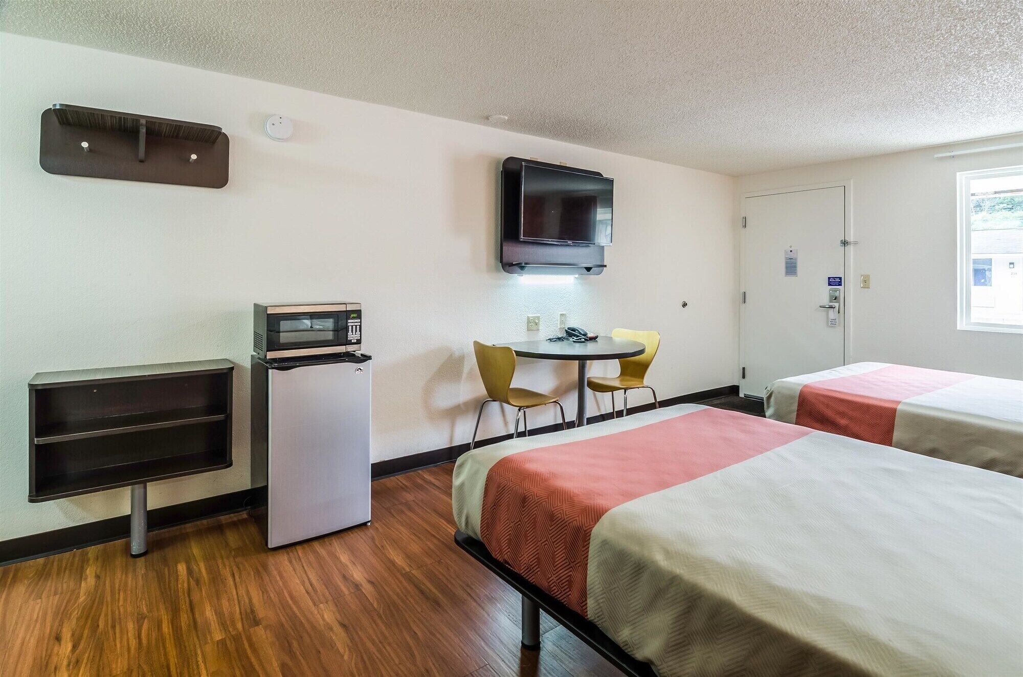 A hotel room with two single beds, a small kitchenette with a microwave, a refrigerator, a TV mounted on the wall, and a small table with two chairs.