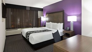 Habitación de hotel con una cama grande, un escritorio, una silla, una mesita de noche con lámpara y una ventana con cortinas.