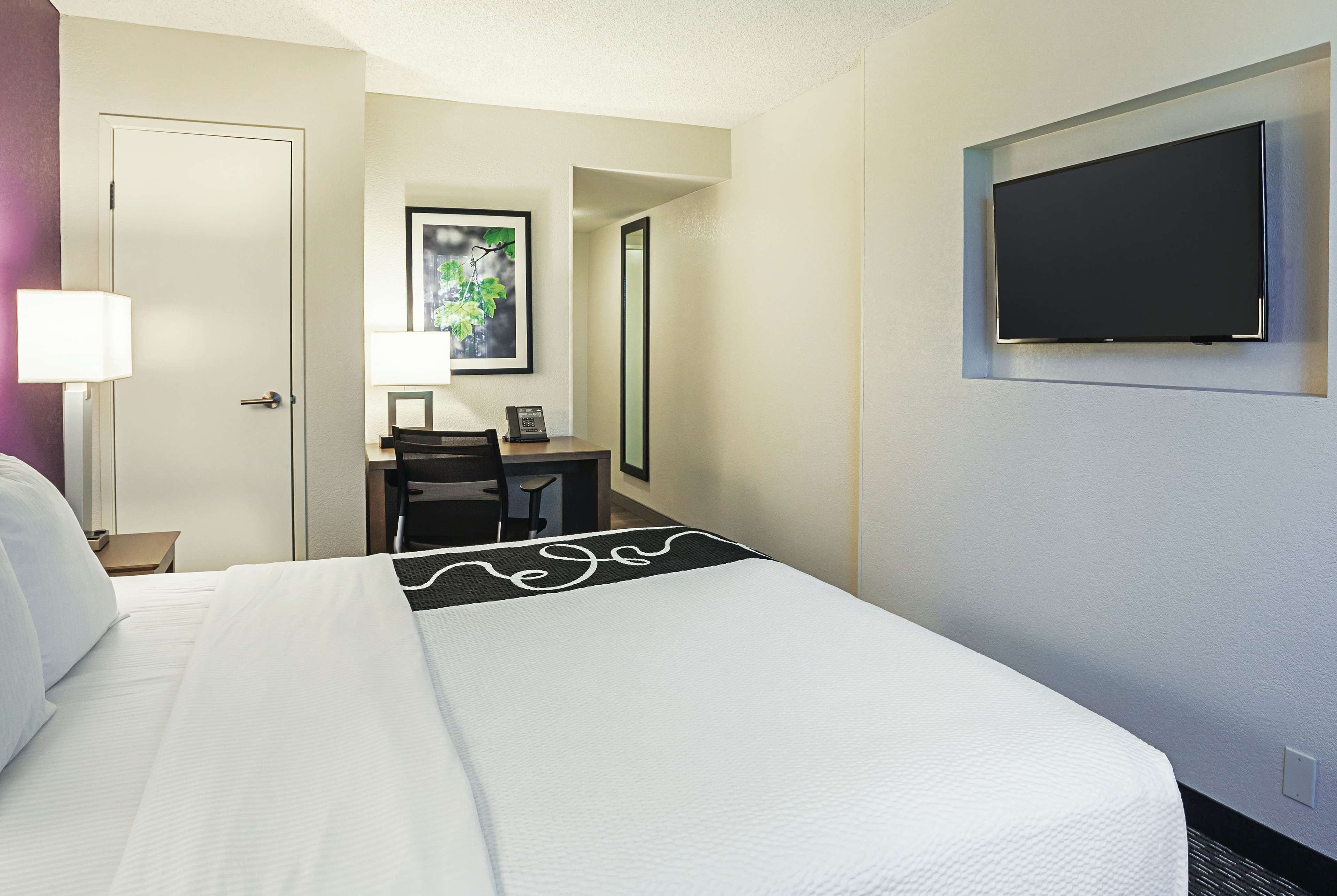 A hotel room with a bed, a desk, a chair, a lamp, a flat-screen TV, and a framed picture on the wall.