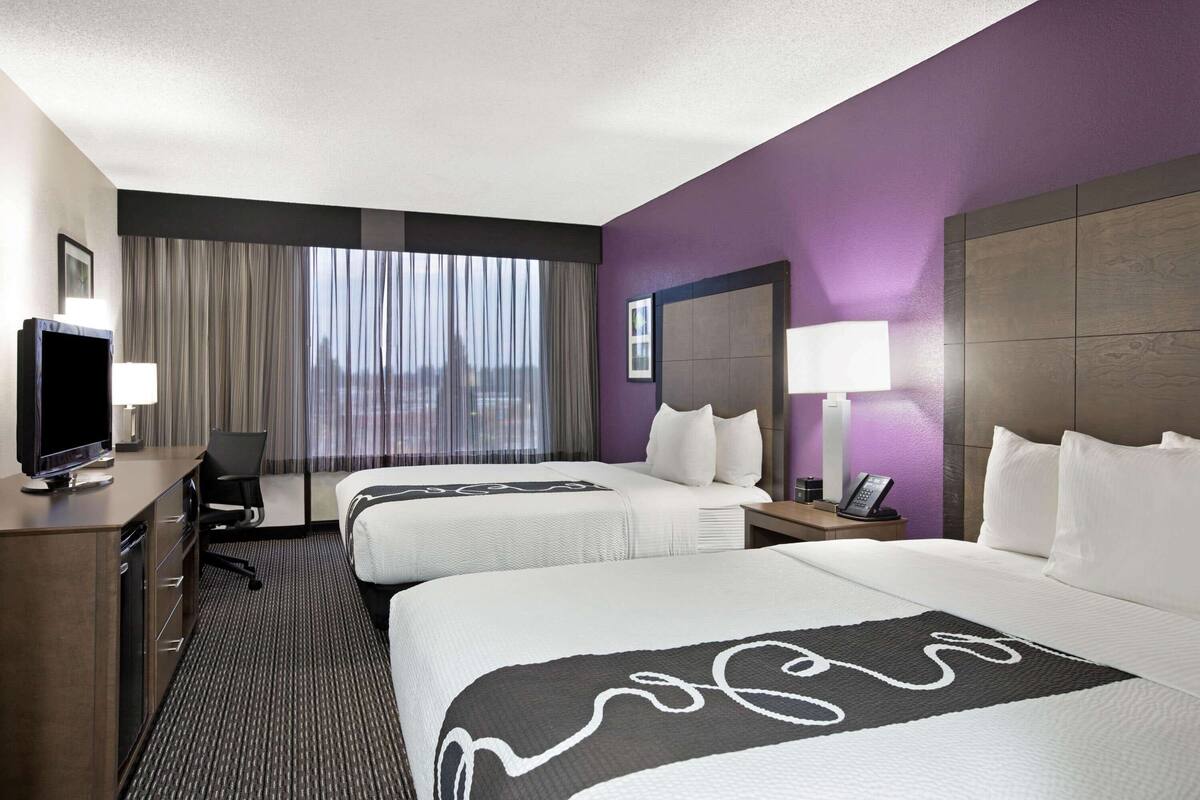 A hotel room with two beds, a desk with a TV, a chair, a nightstand with a phone, and a view of the city through large windows.