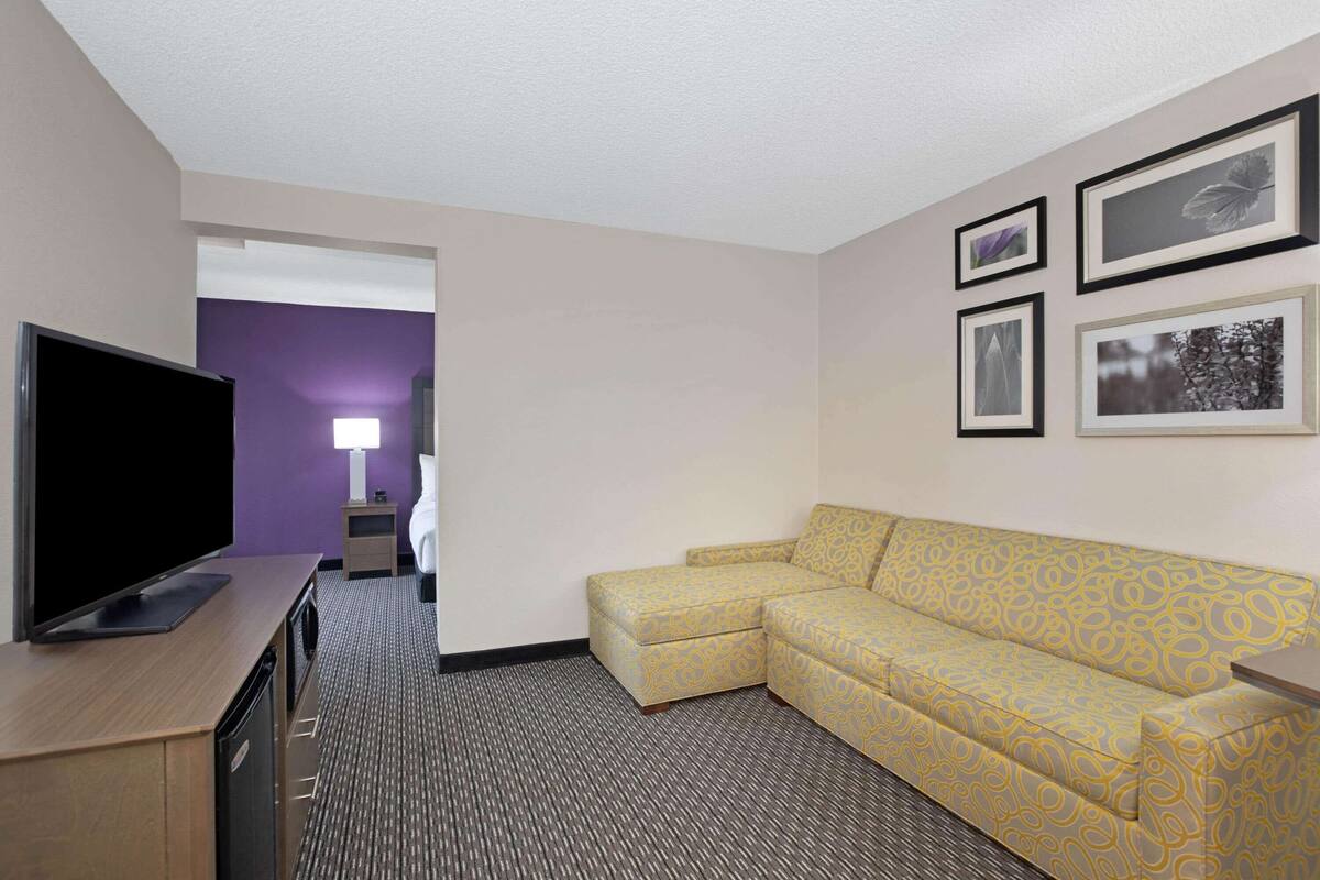 A hotel room with a flat-screen TV on a wooden cabinet, a yellow patterned sofa, a patterned carpet, and framed pictures on the wall.