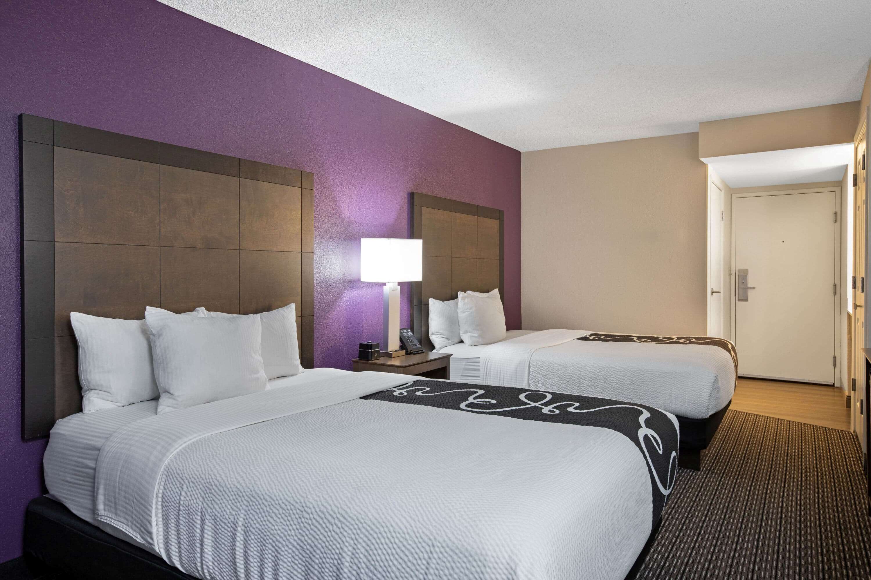 A hotel room with two beds, a nightstand, a lamp, and a door leading to another room.