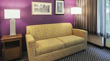A room with a purple wall, a yellow patterned sofa, two bedside tables with lamps, and framed pictures on the wall.